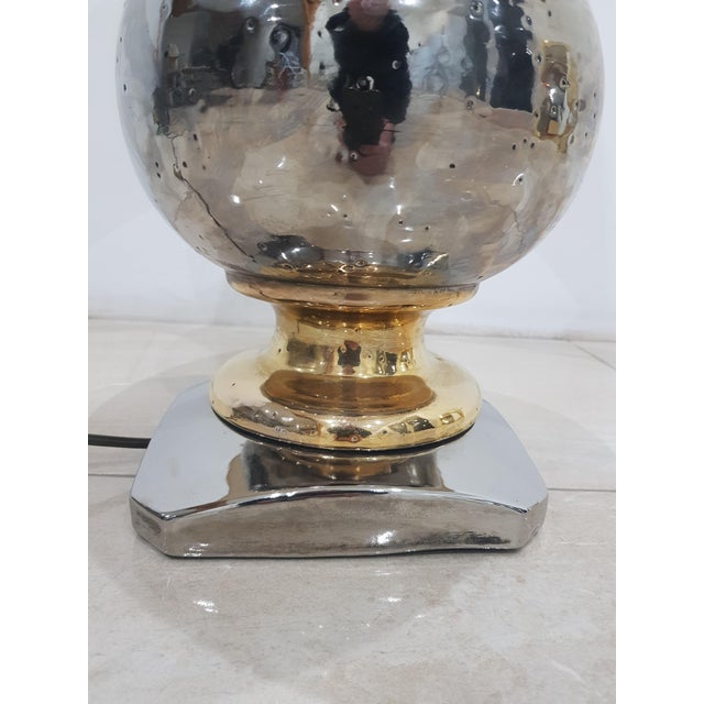 A spherical lamp made of ceramics covered with shiny silver and gold glaze. The lamp stands on a silver, square base with...