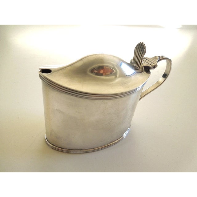 An oval shaped body with a scroll-form handle. Domed lid with slot for a spoon. Cobalt-blue glass liner. Hallmarked on the...