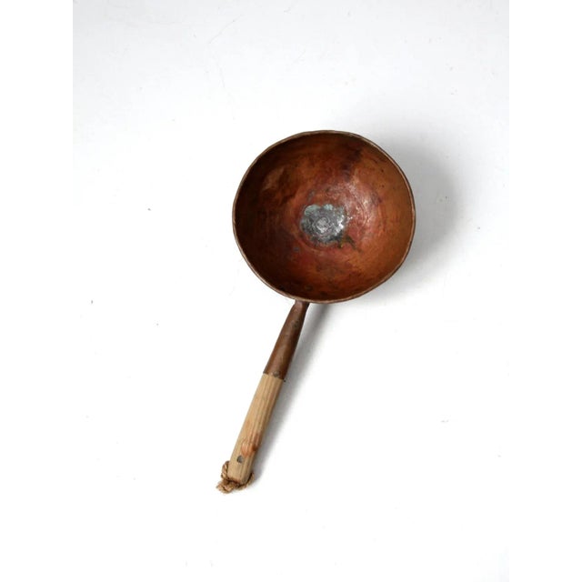 An antique copper pan featuring a rounded base, a wooden end handle, and a rope loop for hanging and storage. The warm...