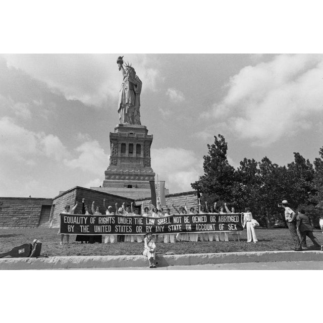 ERA Protest American activist Isolda Dubic sits in front of a group of women standing in front of the Statue of Liberty...