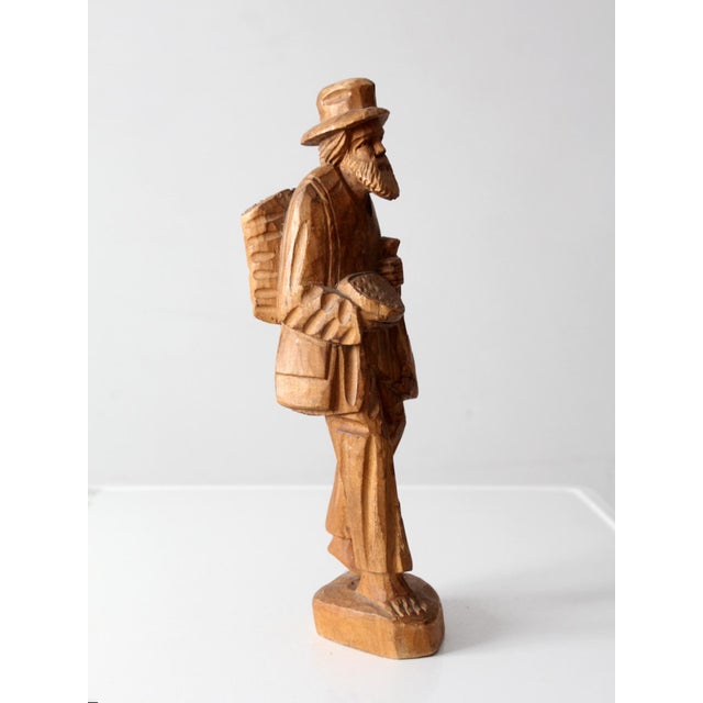 Hand-carved wood figure of a traveling peddler, rendered with expressive, faceted carving and a warm, natural tone. The...