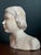A gentle, observant portrait of a young woman, modeled in plaster and finished by hand. The sculptor’s touch is present...