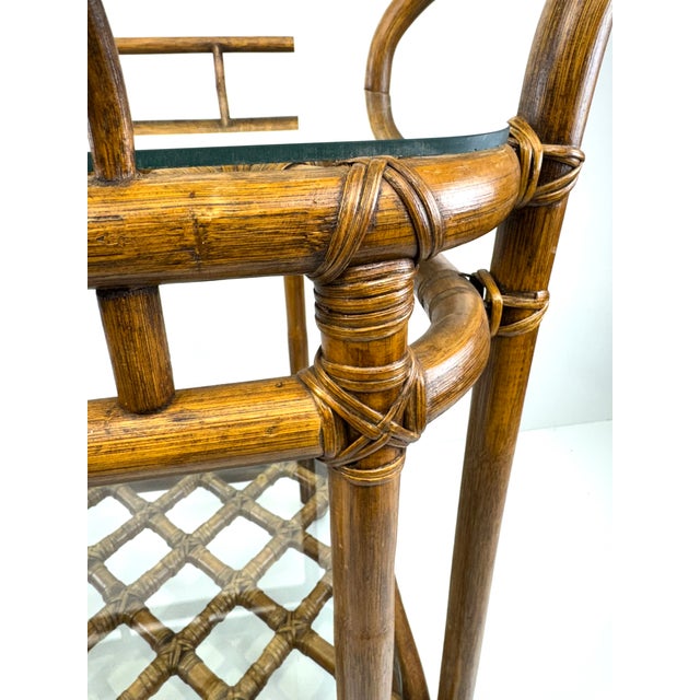 A sophisticated mid-century rattan bar cart in the style of McGuire, featuring two tiers of inset glass shelves framed in...