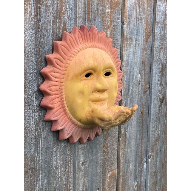 This unique piece of outdoor decor is a terracotta sun face wall hanging, specifically featuring cupped hands protruding...