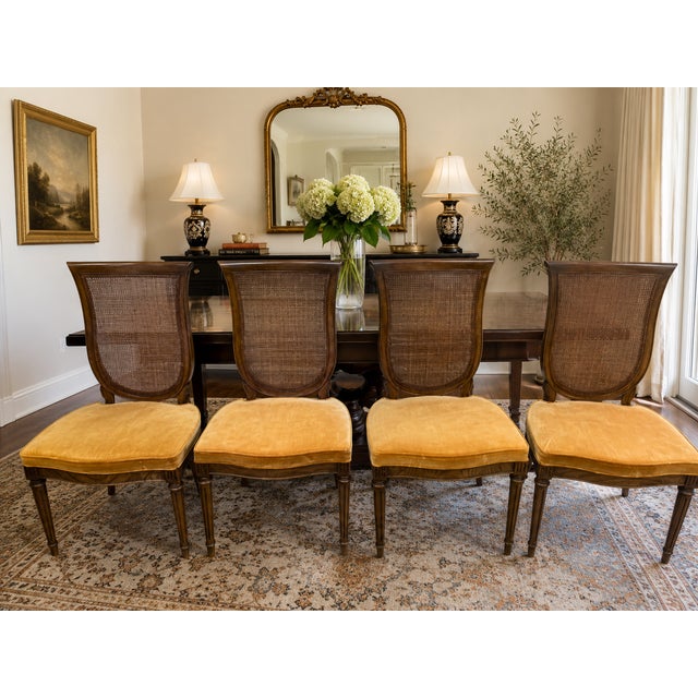 A handsome set of four vintage Neoclassical dining chairs featuring elegant cane backs, warm walnut-toned wood frames, and...