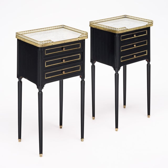 An elegant pair of French Louis XVI style side tables or nightstands featuring ebonized wood cases, brass detailing and...