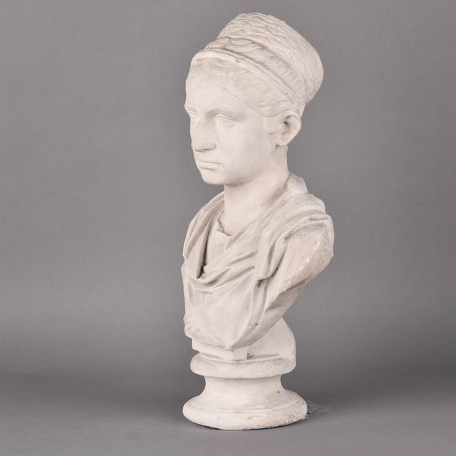 This plaster bust depicts a woman in the classical manner, her hair drawn back beneath a wrapped fillet into a high coiled...