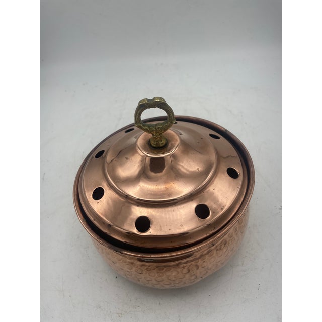 Lidded copper pot with perforated top and a round handle. The pot has a rolled rim and rounded body. The perforations in...