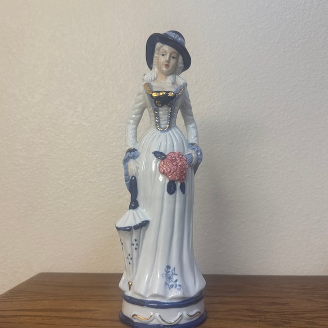 An elegant vintage porcelain figurine depicting a stylish woman in historical dress. Standing gracefully, she wears an...