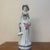 An elegant vintage porcelain figurine depicting a stylish woman in historical dress. Standing gracefully, she wears an...