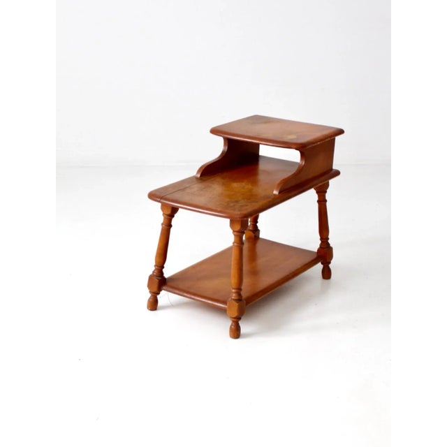 A mid-century wooden end table from around 1950, showcasing a casual ranch-style design. The table features a tiered top...