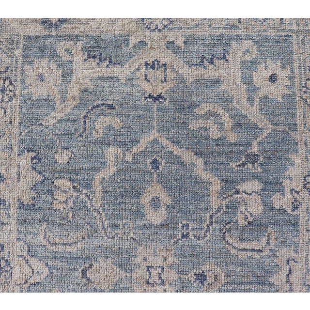This Oushak Runner is rendered in a medium blue background and a light grey border. The design is an arabesque-like floral...
