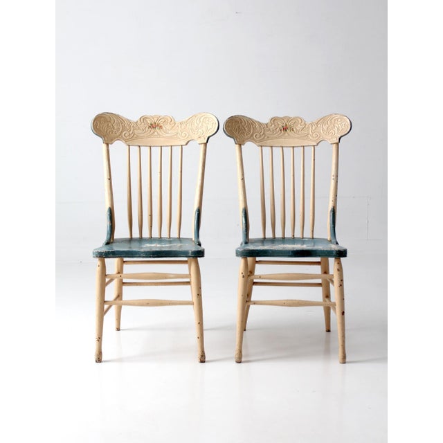 This charming pair of antique painted spindle back chairs features carved crest rails, floral detailing, and worn blue-...