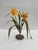This artwork features a delicate arrangement of yellow flowers, crafted from paper and paint, emerging from a textured...