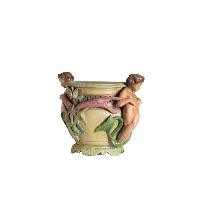 A polychrome ceramic planter features an ornate sculptural design with cherubs in high relief, intertwined with botanic...
