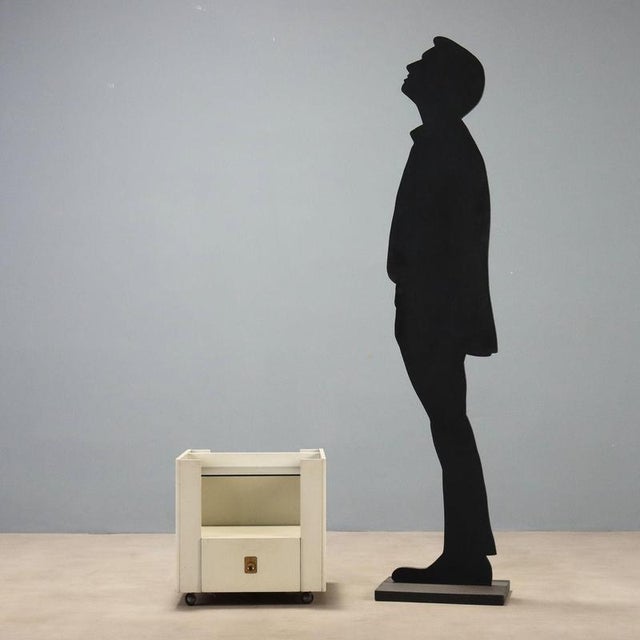 1970s nightstand crafted from white lacquered wood, featuring a simple and functional design typical of the era. The piece...