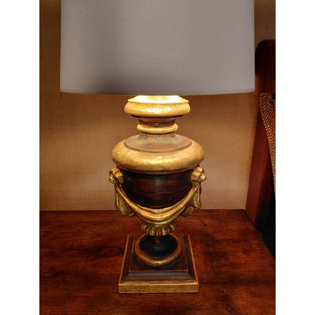 An elegant neoclassical-style table lamp crafted of carved wood with a rich black and deep mahogany-toned finish. Gilt...
