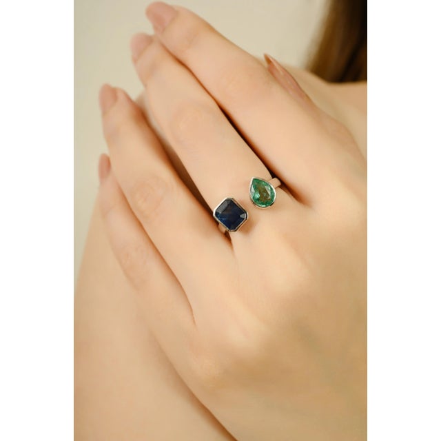 This ring is crafted in 18k white gold, featuring an emerald-cut blue sapphire and a pear-cut emerald, together weighing...