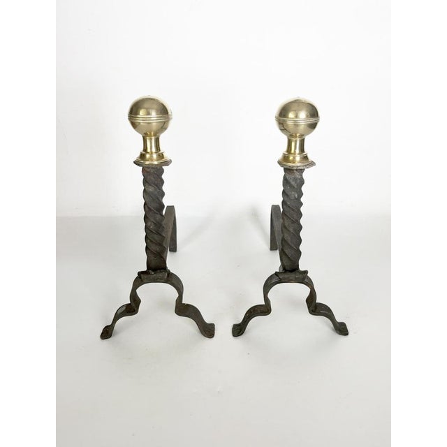 An elegant pair of fireplace andirons combining a black cast-iron structure with a polished brass decorative sphere. The...