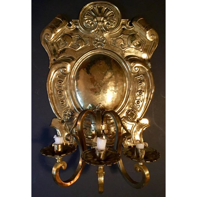 This is an impressively large and ornate antique repousse brass three-light sconce. The backplate is crest shaped with a...