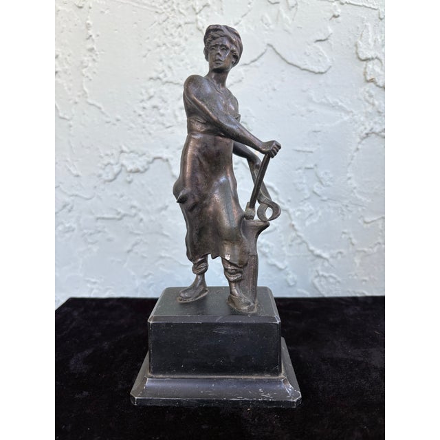 This early 20th century figurative sculpture depicts a blacksmith, captured in a moment of quiet pause while holding his...