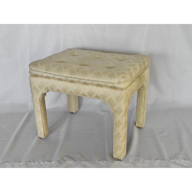 An upholstered low stool. The square leg with faux bracket supports the button tufted cushion top. In the style of Billy...