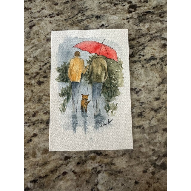 A tender moment captured in soft watercolor, “Under One Red Umbrella” depicts two figures walking arm in arm beneath a...