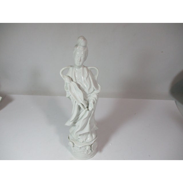 White porcelain female Asian figurine in a calming pose. She is dressed in traditional flowing robes, holding a large...
