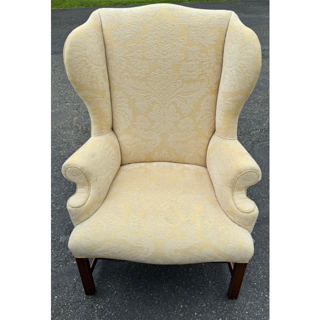 Generously proportioned wingback chair with pale yellow damask upholstery, block legs with stretcher, rolled arms and...
