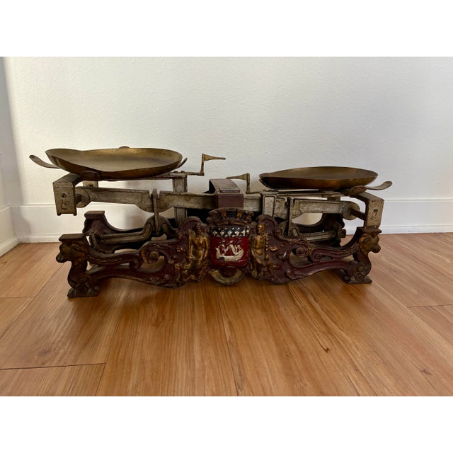 Large antique balance scale featuring twin brass pans mounted on an ornate cast metal base with sculptural detailing and a...