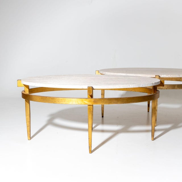 Two oval coffee tables crafted from gold-patinated metal featuring travertine tabletops. These tables are supported by...
