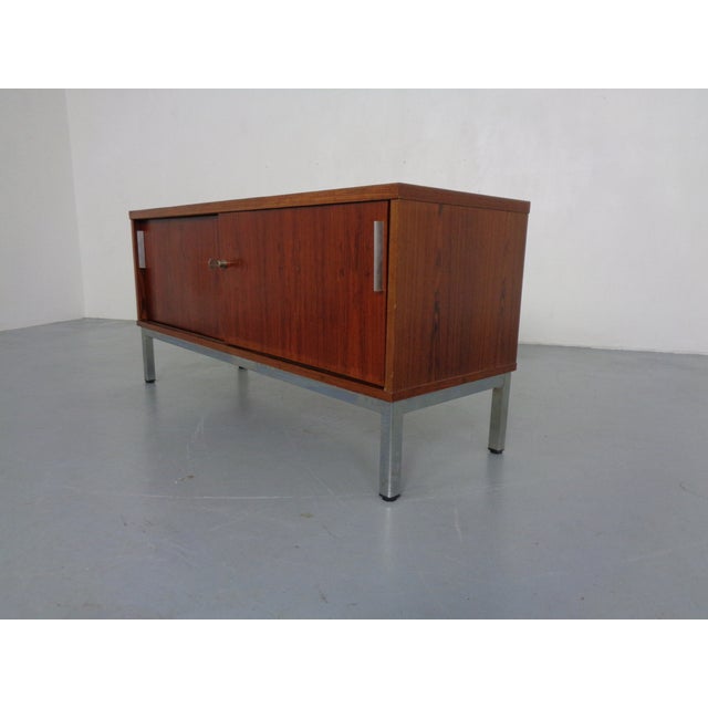 A small, rosewood-veneered sideboard from the 1960s, likely made in Germany. It features two sliding doors, each...