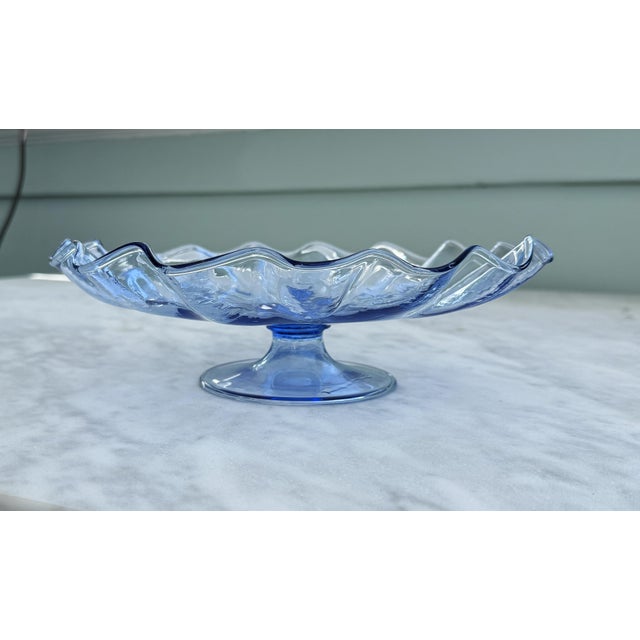A beautiful mid‑century hand‑blown art glass pedestal bowl in a soft light‑blue tone, featuring a scalloped rim, natural...