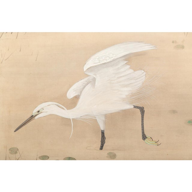 This serene Meiji-period Japanese two-panel screen depicts a graceful white heron standing at the edge of a tranquil pond...