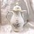 This is a Rosenthal Coffee Pot in the Grey Rose pattern from 1971. The pot is white with a woven design, gray and pink...
