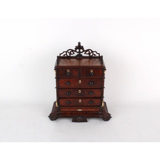 A miniature mahogany chest of drawers in a rich reddish-brown wood grain finish with a pierced, scrolling foliate gallery...