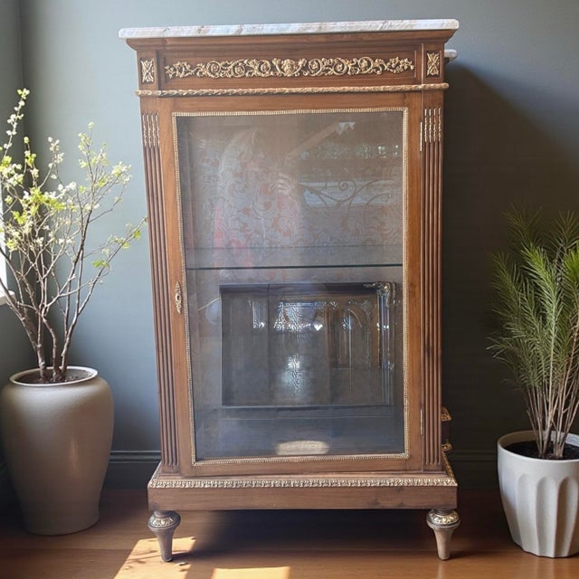 French Louis XVI Marble-Top Display Vitrine • Gilt Carved Accents • Glass Curio Cabinet (2 Available) Description- A...