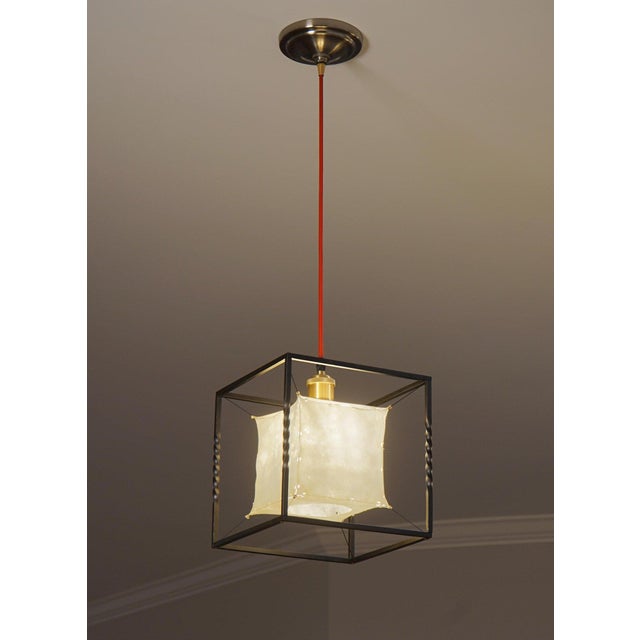 LIGHTMASS P1 is a handmade pendant light with a natural sheepskin shade suspended inside a cube frame of heat-blackened...