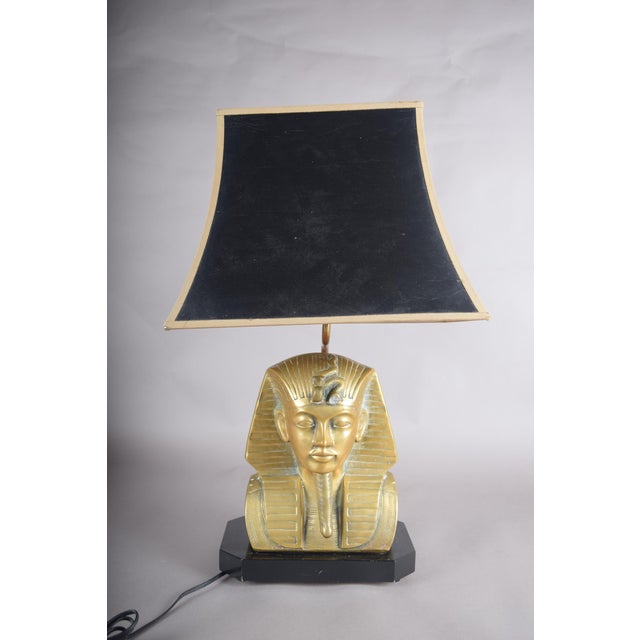 A bold and iconic vintage Egyptian Revival table lamp featuring a striking pharaoh bust in a rich brass tone, set on a...