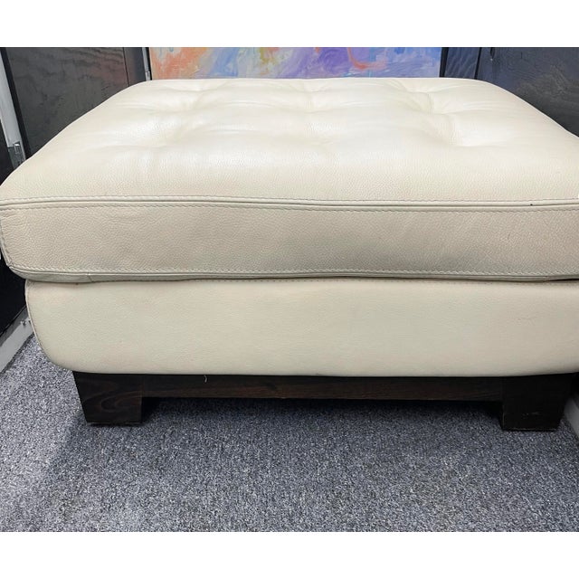 Roche Bobois square ottoman in cream pebbled leather with button tufting and a solid dark wood plinth base. A contemporary...