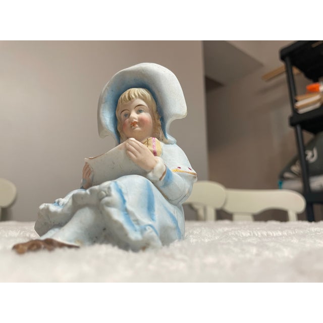 Vintage bisque porcelain figurine depicting a young girl wearing a soft blue bonnet and coordinating dress. This charming...