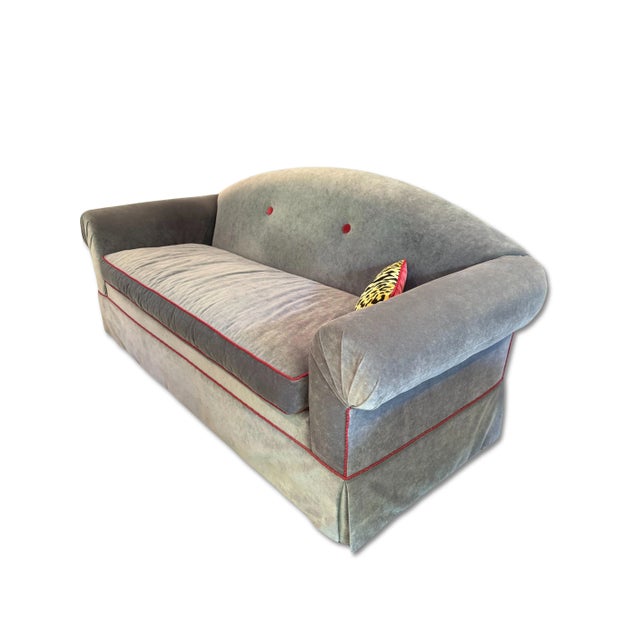 Camelback sofa in gray velvet, designed with a single bench cushion, a skirted base, and accented with contrasting red...