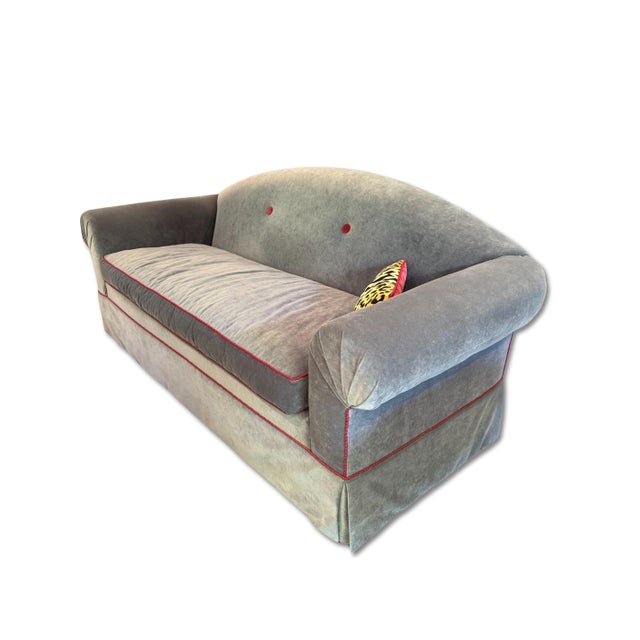 Camelback sofa in gray velvet, designed with a single bench cushion, a skirted base, and accented with contrasting red...