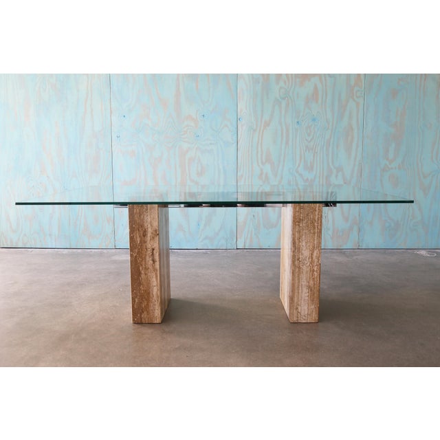 A postmodern dining table attributed to Artedi. Travertine bases linked with a chrome bar supporting a glass top. This...