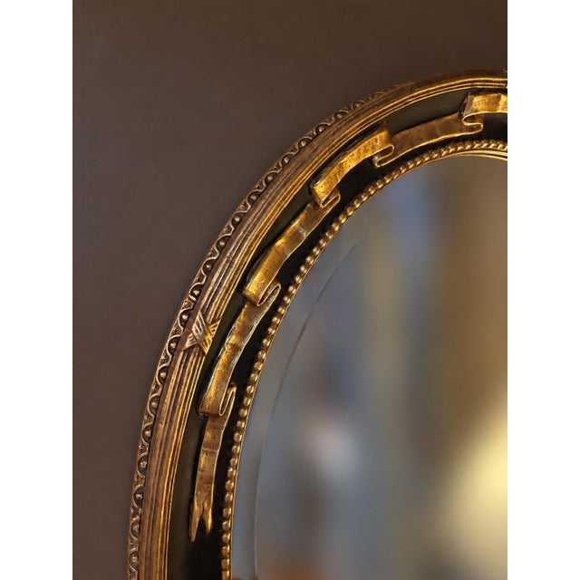 Elegant vintage Federal Revival oval wall mirror featuring a dramatic black and parcel-gilt frame. Raised gold ribbon and...