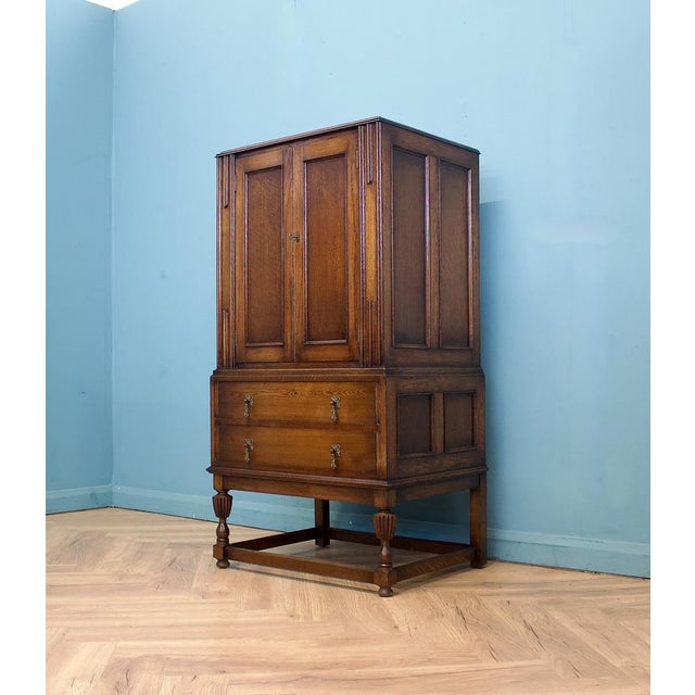 A solid oak cabinet dating to the 1940s, with a warm tone and simple panelled doors. The upper section opens to two glass...