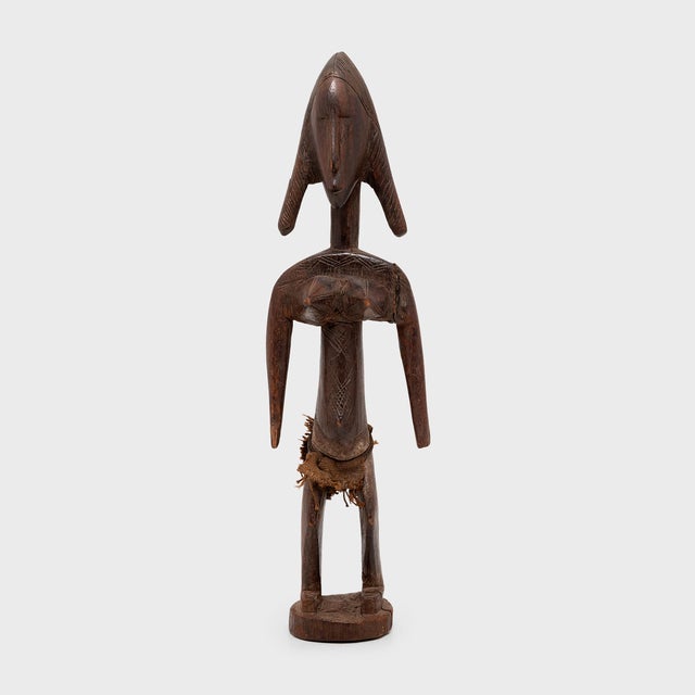 With angular curves, refined facial features, and plaited hair, this wooden figure by the Bambara people of Mali is carved...