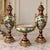 Elegant three-piece French-style porcelain garniture set featuring two matching covered vases and a coordinating central...