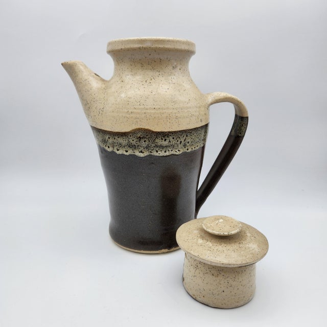 Hand-thrown stoneware lidded pitcher with speckled cream upper glaze, dark brown lower half, and a textured transitional...