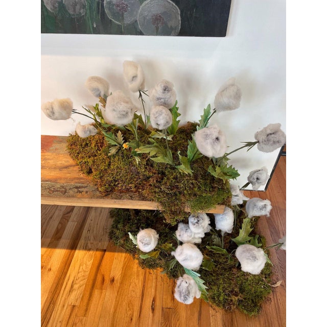 Wool Over My Eyes features a whimsical arrangement of soft, woolen forms resembling flowers, emerging from a lush, mossy...